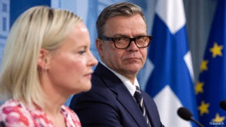 Riikka Purra ja Petteri Orpo lehdistötilaisuudessa Suomen ja EU:n lippujen edessä; taustalla näkyy mikrofonit, ja ilmapiiri on virallisen lehdistötilaisuuden mukainen.